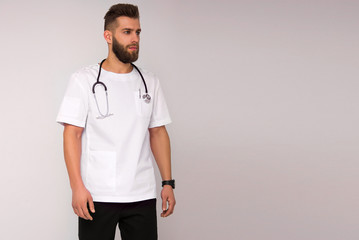 young stylish nurse male in white medical shirt and black pants with stethoscope on his neck is standing and looking straight on white wall background. medical concept. free space on right side