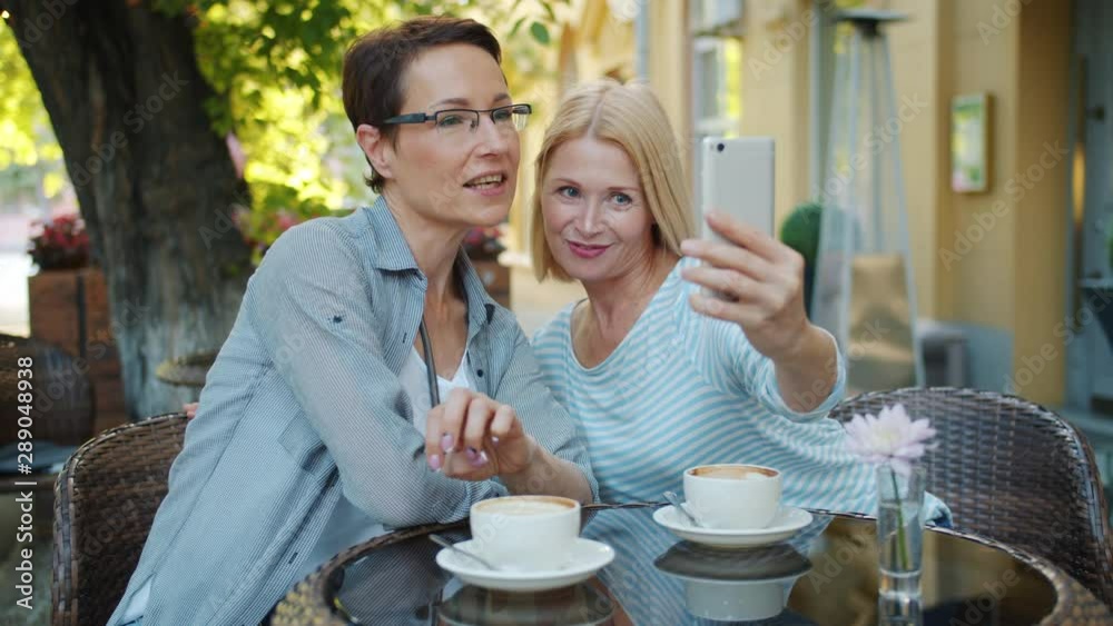 Wall mural Beautiful mature women friends are taking selfie with smartphone camera and talking posing in outdoor cafe. Photograph, people and modern lifestyle concept.