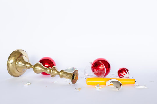 Front view of shattered red and silver Christmas baubles and an overturned bronze candleholder with a yellow candle on white background