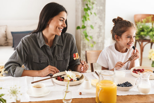 Image of joyous family mother and little daughter eating tasty food together while having breakfast at home in morning