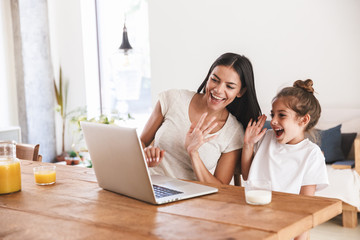 Obraz premium Image of caucasian family woman and her little daughter smiling and waving at laptop computer together in apartment