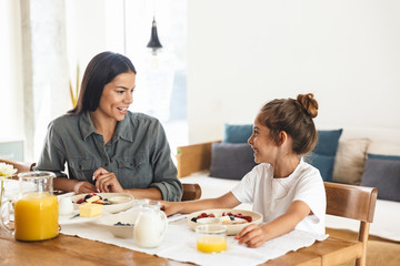 Image of positive family mother and little daughter having breakfast at home in morning