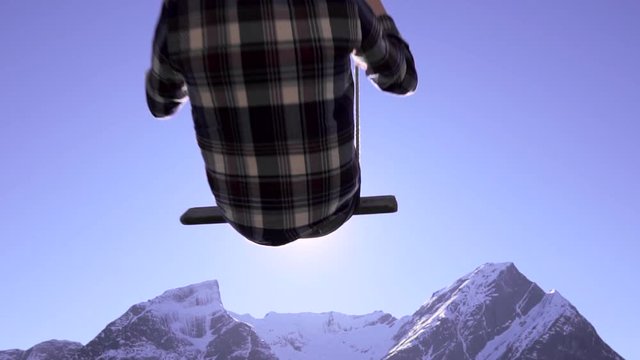 Slow motion shot of person riding on a swing, flying towards the sun, beautiful scenic view of snow-covered mountains.