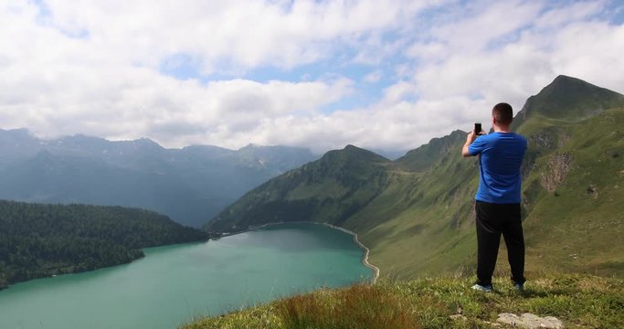 Pan right to left, showing a man taking a photo on top of a mountain, looking down towards Lake Ritom.