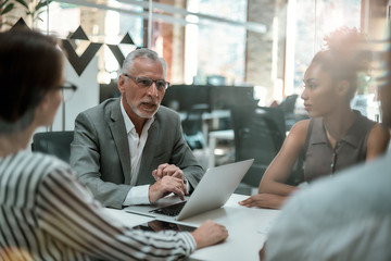 Brainstorming. Serious mature man in formal wear discussing something with his multicultural team while sitting at the office table