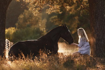 portrait of beautiful young woman with blond hair sitting in front of lying horse and feed it from...