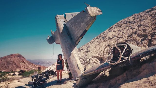 4K UHD Cinemagraph of a young brunette woman standing next to a wreck of an old American military air plane. Her hair is gently moving in the wind.