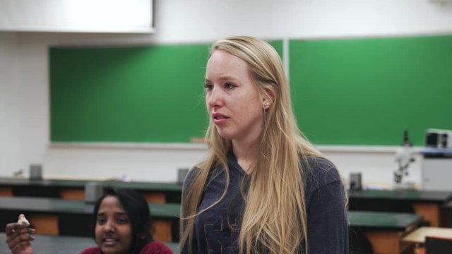 An attractive young blond-haired student is standing in a classroom and makes a short smile to finally be serious again