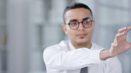 Chest-up shot of Arab man in white shirt, tie and glasses using invisible interactive augmented reality touchscreen, with rotating, moving and tapping gestures. Suitable for adding AR, VR elements.Che