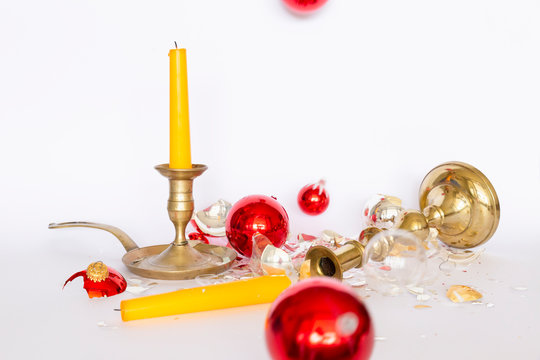Front view of falling and shattered red and silver Christmas baubles and two bronze candleholders with a yellow candle on white background