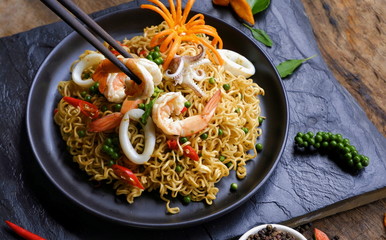 Close up chopstick pickup shrimp with stir fry instant noodles spicy seafood in black plate on slate and wooden table floor background