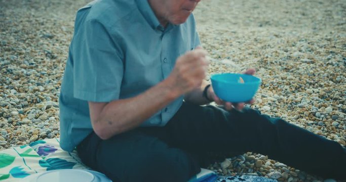 Senior man eating salad and sausages on the beach in summer