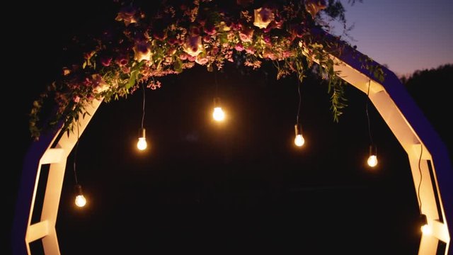 Beautiful wedding decorations arranged outdoor in night park. Rounded wooden frame decorated with fresh roses, strings with glowing electric bulbs and burning candles.