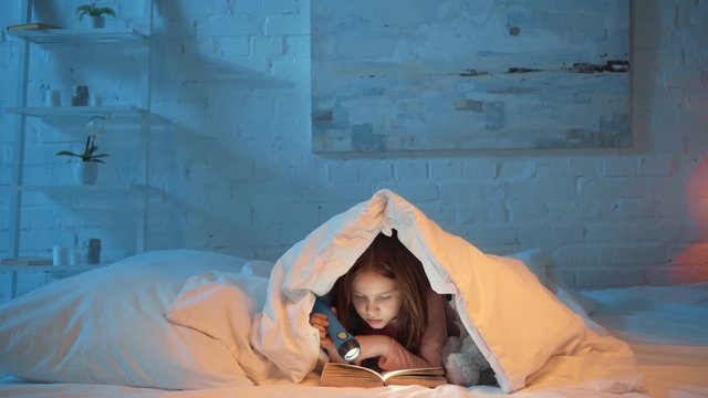 scared kid under blanket reading book with flashlight at night