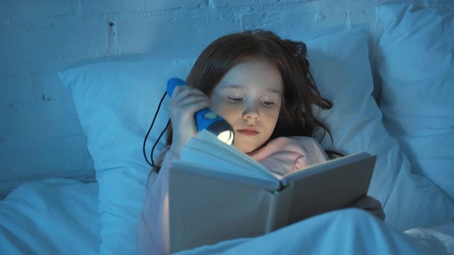 emotional child reading book with flashlight in bed at night