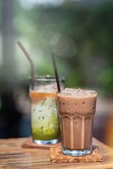 Vertical shot of iced mocha and iced matcha tea in glasses with straws on wooden table outdoors. Perfect beverages for a hot summer day. Selective focus