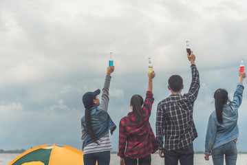 Evening at the beach. Group of friends are drinking beers and having fun, sitting in the sand around a campfire. They are smiling doing toast and clinking their beers. The men are shirtless.