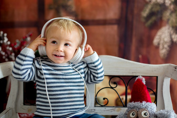 Sweet toddler boy with headphones, listening to music, sitting on rustic bench, christmas...