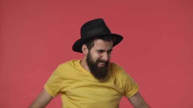 Angry devastated middle-eastern jewish guy with beard in black hat, yellow t-shirt, clenching fists in anger gesturing outraged and upset, feeling disappointment from failure, stand red background