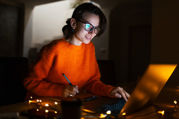 Portrait of smiling designer girl, working at graphic tablet on laptop. Wearing eyeglasses and orange sweater. In dark room home, with garlands.