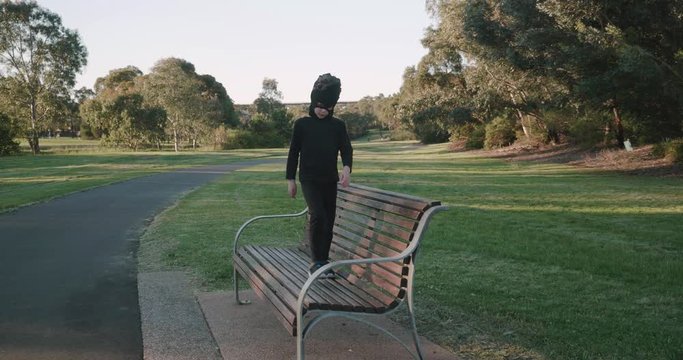 young boy wearing a homemade ninja mask and black costume climbs on to a park bench and then jumps off then walks towards the camera  in a park in the late afternoon golden glow of the setting sun
