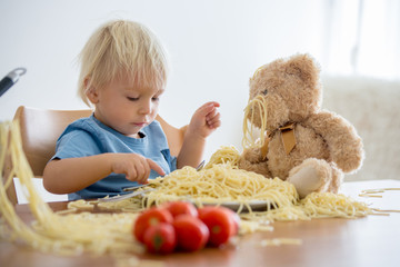 Little baby boy, toddler child, eating spaghetti for lunch and making feeding teddy bear friend