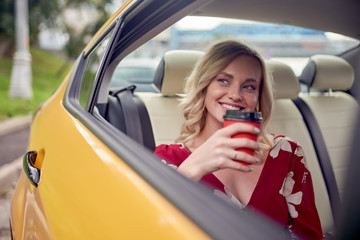 Photo of happy blonde with glass of coffee in her hands sitting in back seat of yellow taxi