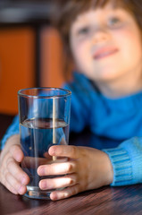 Adorable smiling little girl holding glass of water in kitchen. Glass is focused, blurred girl. Health and beauty concept