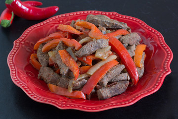 Cooked liver with vegetables on red plate on black background