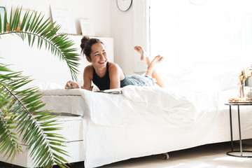 Portrait of pretty woman smiling while lying and reading magazine on bed in bright room with green...