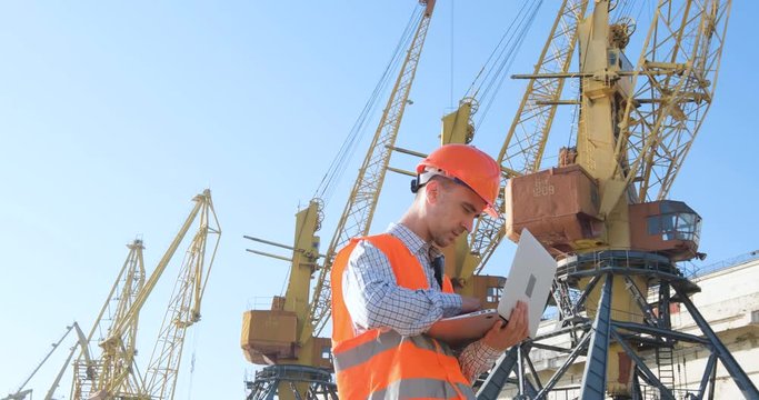 male worker of sea harbor in orange helmet and safety west, cranes and sea background 4K UHD futage 	