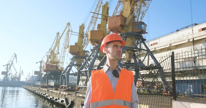 male worker of sea harbor in orange helmet and safety west, cranes and sea background 4K UHD futage 	