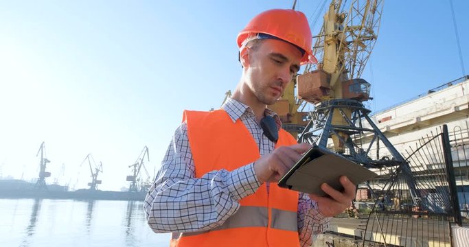 male worker of sea harbor in orange helmet and safety west, cranes and sea background 4K UHD futage 	