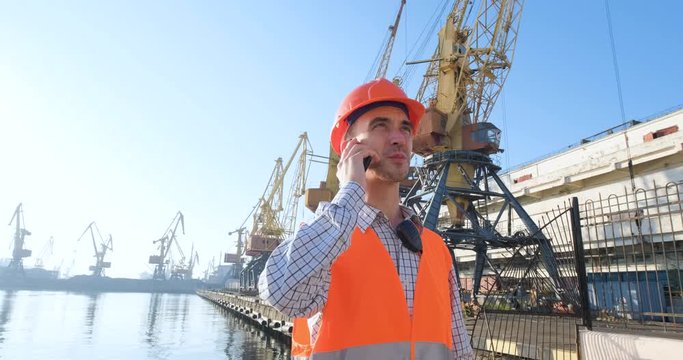 male worker of sea harbor in orange helmet and safety west, cranes and sea background 4K UHD futage 	