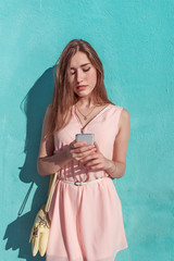 Beautiful girl in pink dress, background turquoise blue wall, in hands of mobile phone, reads and writes a message, online application on wifi social networks. Yellow bag. In summer in city.