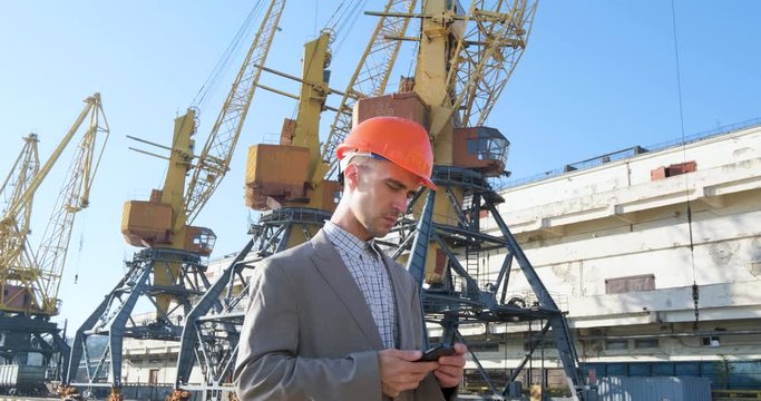 Young male worker of sea harbor in helmet, cargo manager in suit and halmet works outdoor , cranes and sea background	