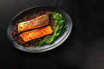 Grilled salmon with sesame seeds, wakame sea vegetable and wild rice, shot from the top on a black background with a place for text