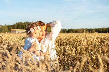 A girl with red hair hugs a little girl, daughter. The family is sitting on the field with ripe ears of corn. Woman and child in white shirts. look at the sunset and rejoice. Countryside and sunset.
