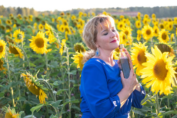 A girl with red hair holds in her hand a glass bottle with sunflower oil. A woman in a blue dress is standing on a field with sunflower flowers at sunset. Copy space