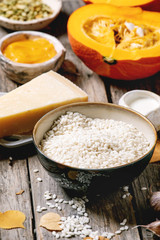 Ingredients for cooking pumpkin risotto. Raw uncooked risotto rice in ceramic bowl, sliced pumpkin, cream, seeds, parmesan cheese, garlic and autumn leaves over dark wooden background. Close up