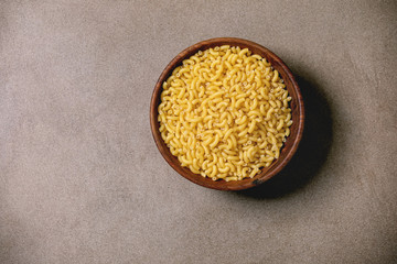 Raw uncooked italian pasta coquillettes in wooden bowl over brown texture background. Flat lay, space. Food background.