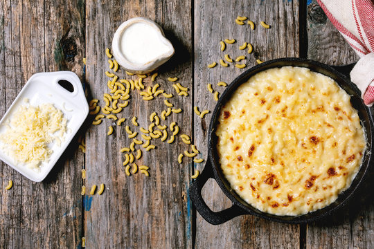 Classic american dish baked mac and cheese in cast iron pan with kitchen towel and ingredients above over old wooden background. Flat lay, space