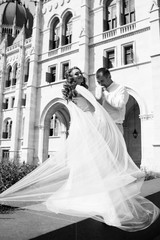 Obraz premium Loving couple together. Attractive young woman in dress and handsome man in suit are stand near building in Prague. The wind waves the dress. Celebrating. Black and white