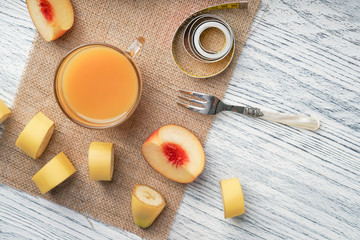 Fototapeta na wymiar A glass of peach juice on a sackcloth on a white wooden table with slices of fruit and a fork and a centimeter