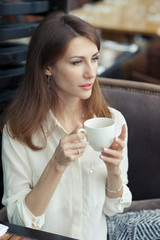 Portrait of a beautiful happy young business woman in a white elegant shirt. girl sitting in a cafe, holding a white cup and looking away