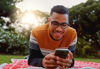 Portrait of an african smiling young man student wearing black eyeglasses relaxing in park using smart phone - college student using mobile in park