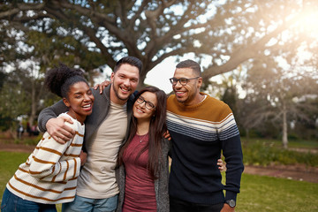 Smiling group of friends laughing together while standing with arms around each other at a park - happy group of people 