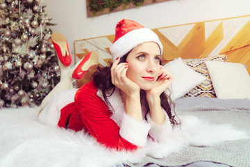young beautiful girl in red Santa Claus suit is lying on the bed and dreamily looking into distance against background of apartment decorated for Christmas..