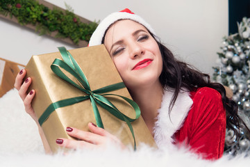 Girl hugs gift lying on the bed. A young woman in the clothes of Santa Claus happy gift for Christmas or new year. Box with a gift in the hands of women