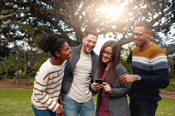 Diverse group of happy friends standing together laughing and looking at smart phone in the park -...